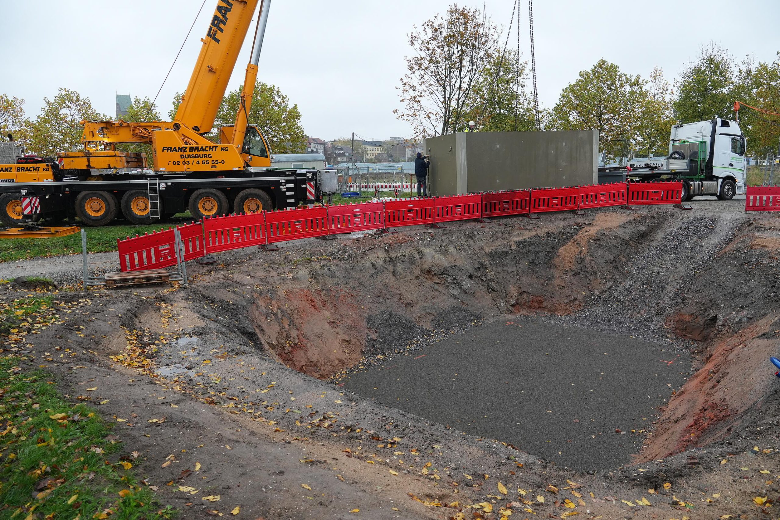 Vorbereitete Baugrube mit betonierter Sohle für die Zisterne, daneben ein Autokran und ein Tieflader mit Betonfertigteil.