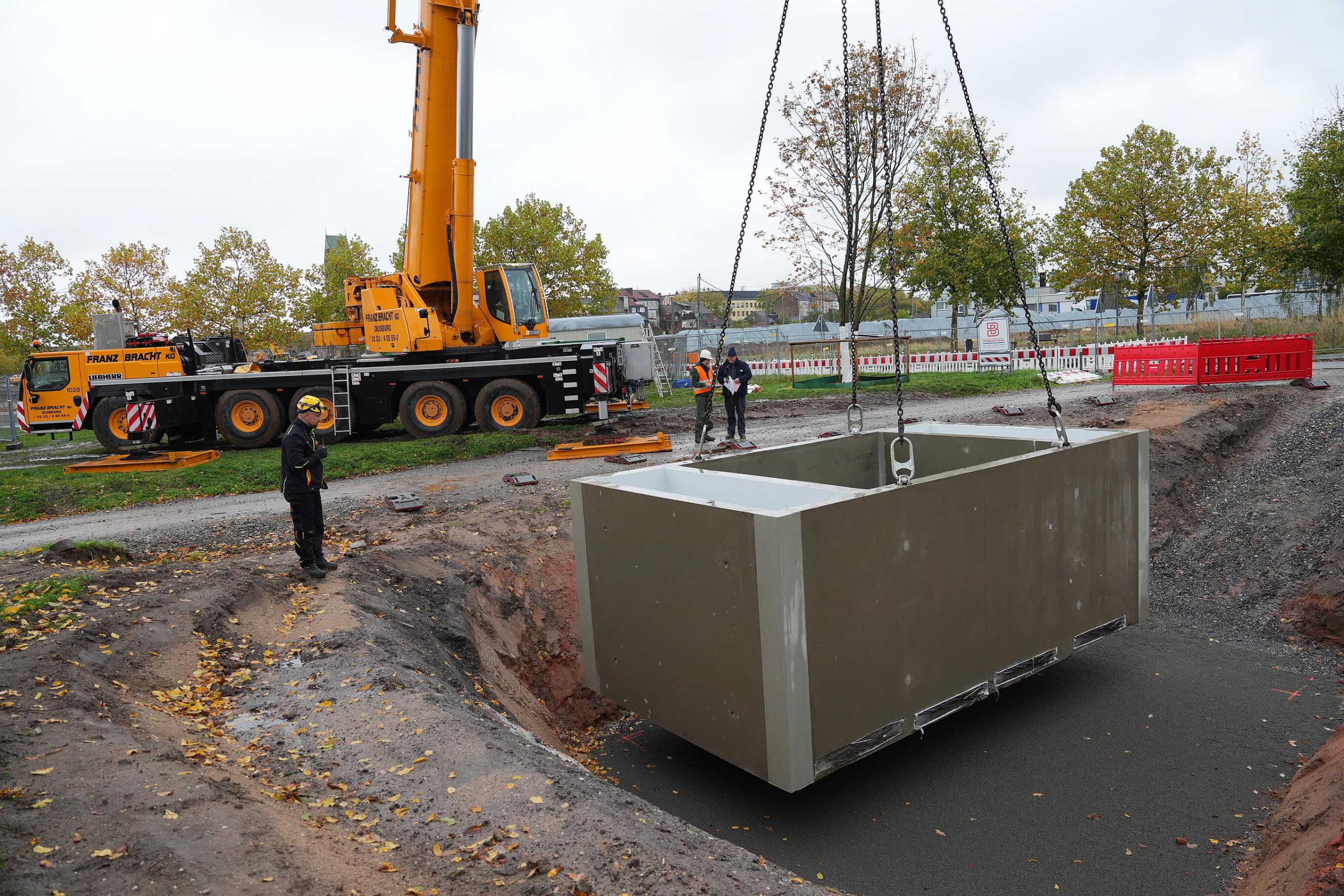 Kran senkt ein großes Betonfertigteil der Zisterne in die Baugrube, Beschäftigte in Warnkleidung begleiten den Einbau