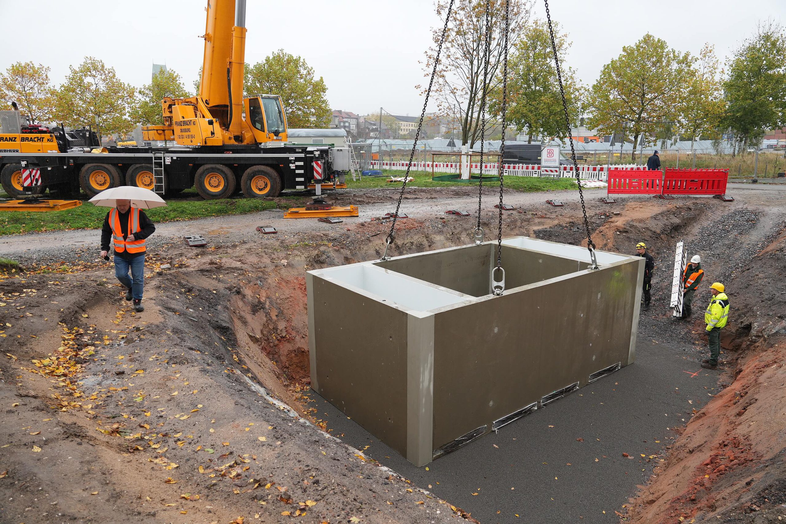 Kran hat ein großes Betonfertigteil der Zisterne in die Baugrube eingesenkt, Beschäftigte in Warnkleidung begleiten den Einbau