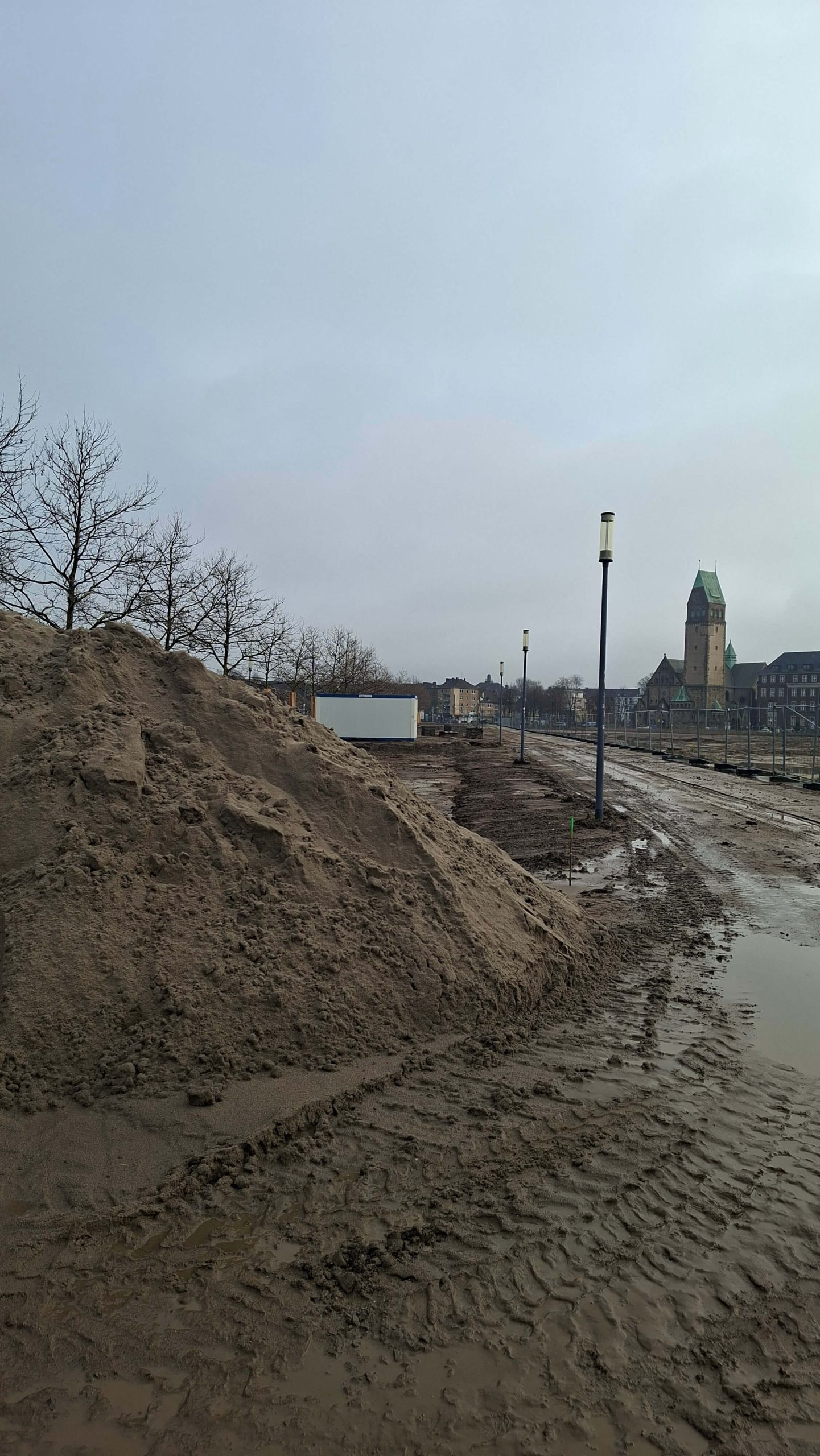 Sandhaufen und aufgeweichter Boden auf der Baustelle der Hochfeldallee, im Hintergrund die Pauluskirche.