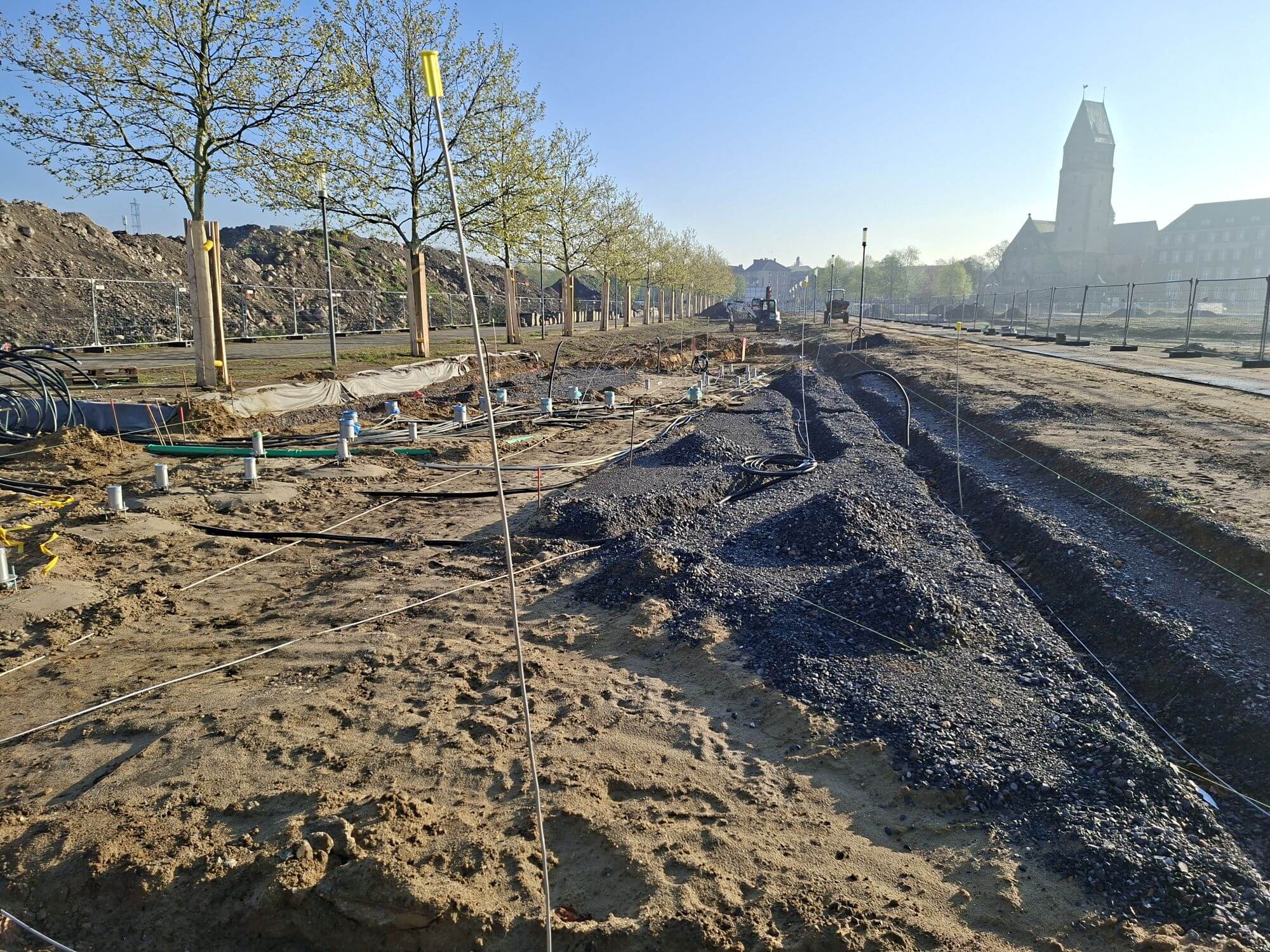 Baustelle am Grünen Ring: Leitungsverlegung und Fundamente entlang einer neu gepflanzten Baumallee, im Hintergrund ein Kirchturm.