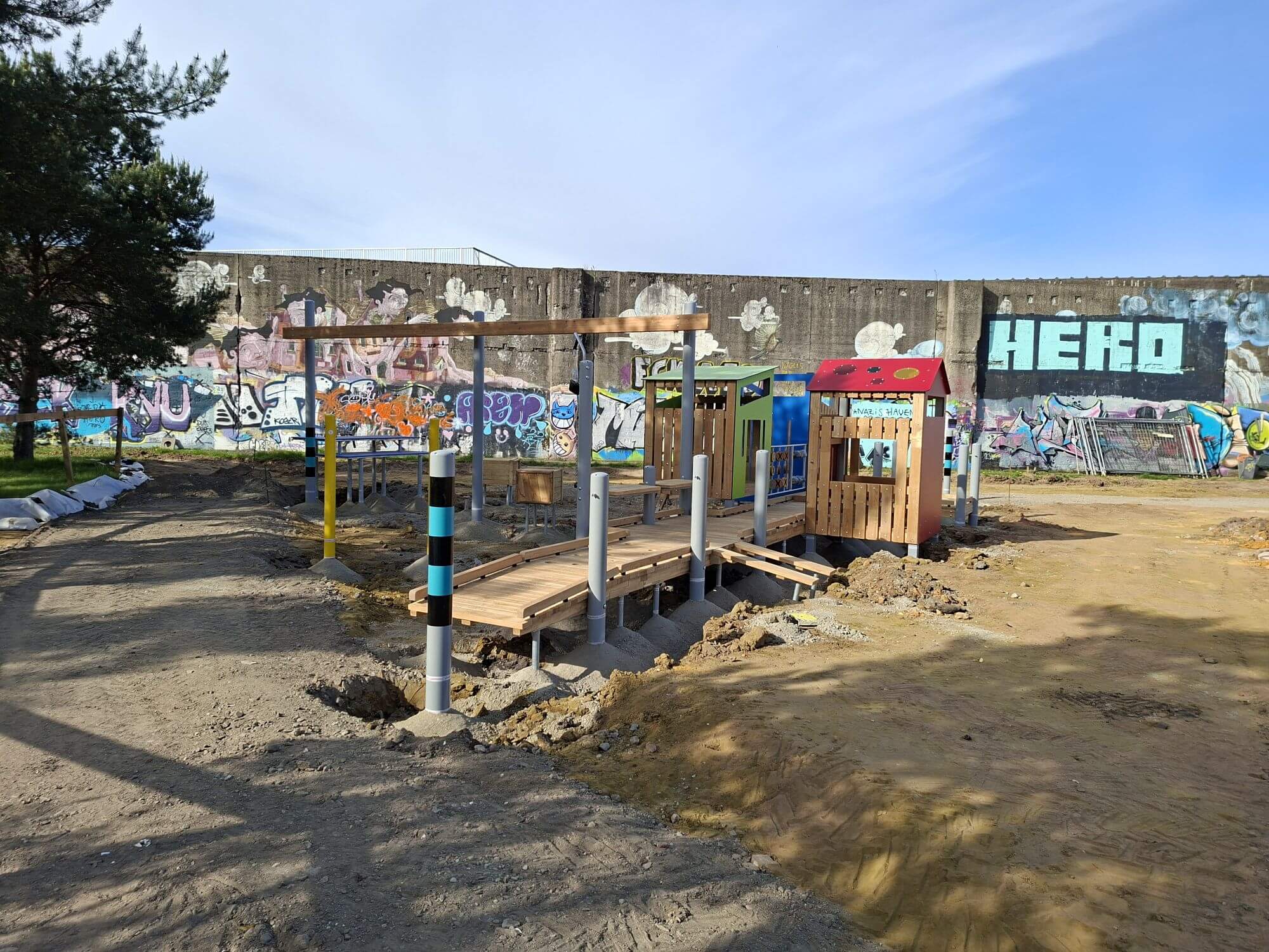 Neu montierte Spielgeräte aus Holz mit Podesten und Spielhäuschen auf dem Spielplatz im RheinPark vor der graffitibesprühten Wand.