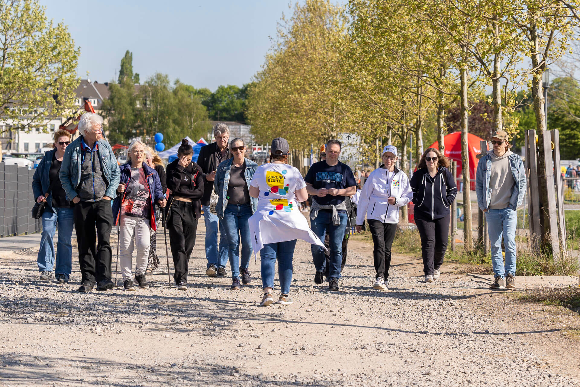 Besuchergruppe spaziert bei Sonnenschein über einen Kiesweg zwischen jungen Bäumen, im Vordergrund eine Frau im weißen T-Shirt mit der Aufschrift „Zukunft. Blüht."