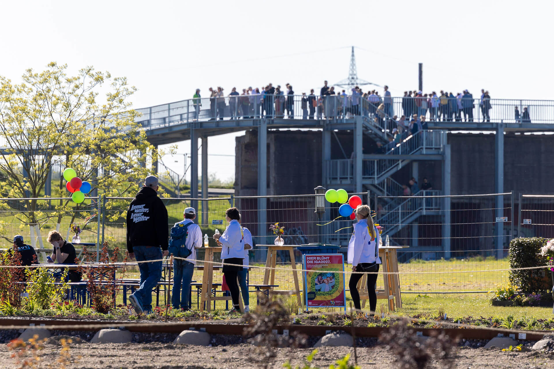 Besucherinnen und Besucher auf dem Skywalk und an Stehtischen darunter beim Tag der IGA im RheinPark, dazu Luftballons und ein Plakat mit der Aufschrift Hier blüht die IGA2027
