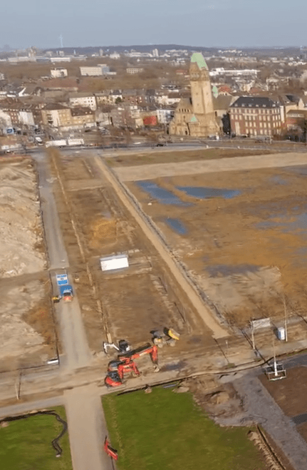 Luftaufnahme der Baustelle der Hochfeldallee: Erdarbeiten mit Baumaschinen und Wasserlachen, im Hintergrund die Bebauung von Duisburg-Hochfeld.