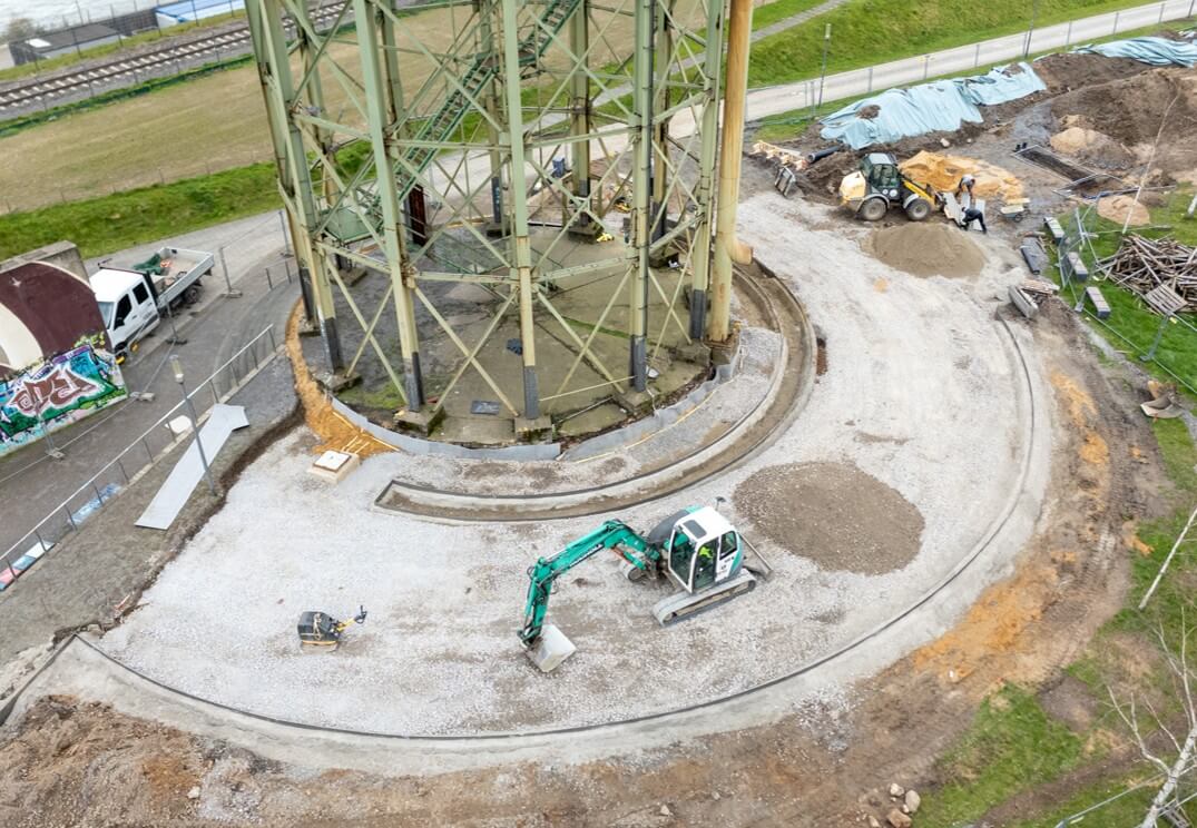 Luftaufnahme der Bauarbeiten am Platz um den Wasserturm: Bagger und Radlader auf der geschotterten Kreisfläche unter dem Stahlgestell.