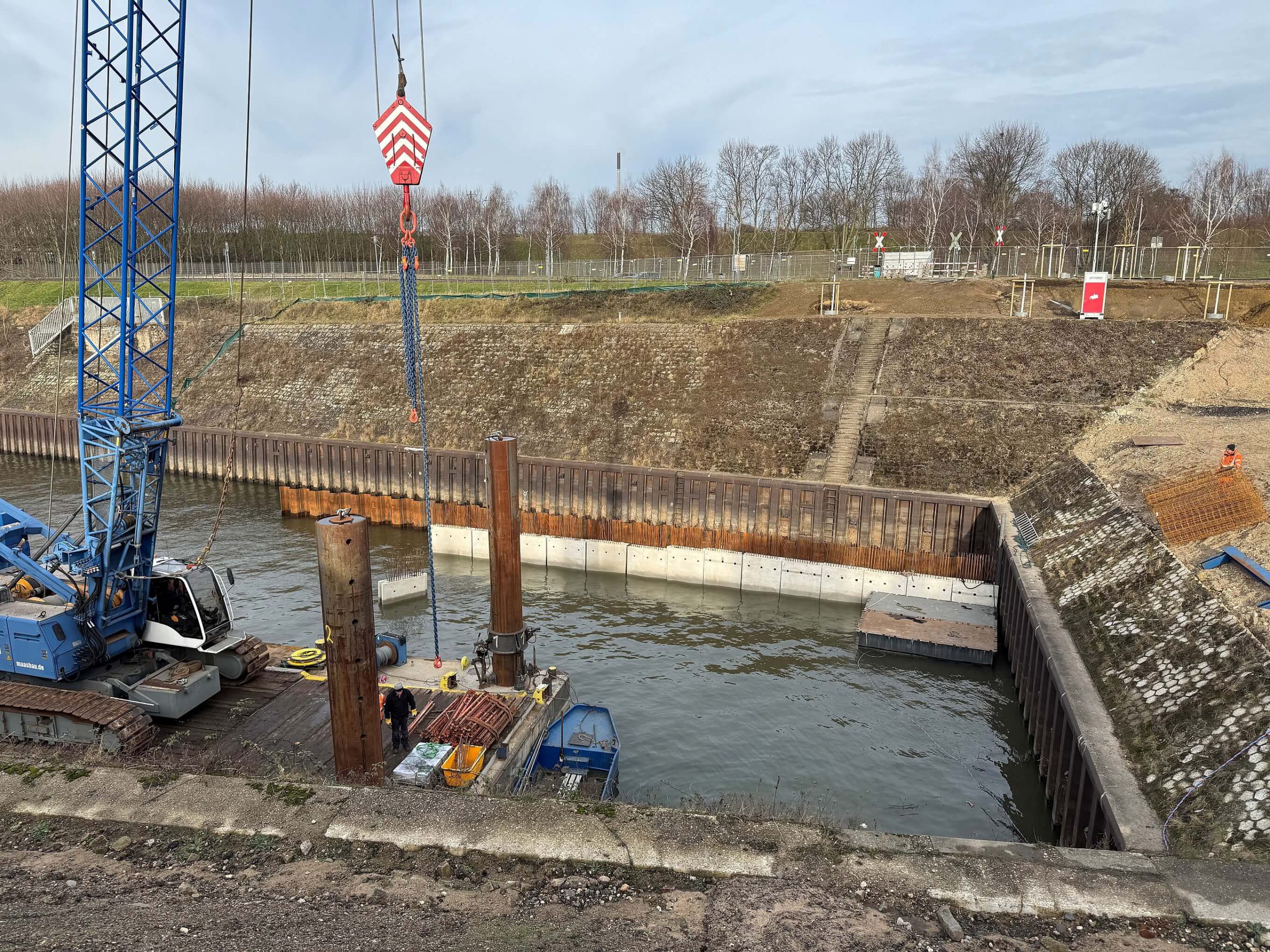 Spundwandarbeiten im Hafenbecken am Kultushafen: blauer Raupenkran auf einem Ponton setzt Stahlprofile, davor die neue Betonwand.