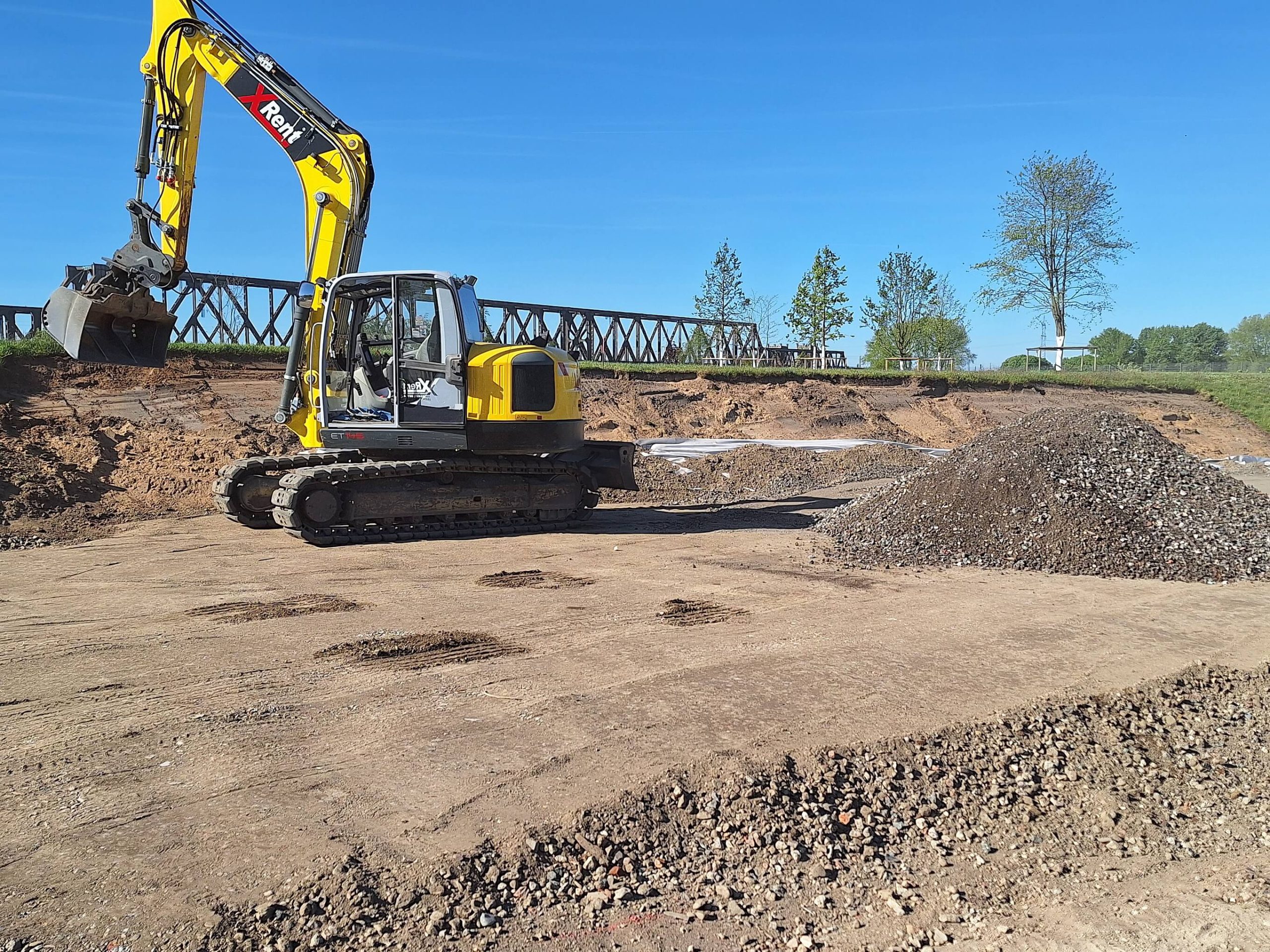 Gelber Bagger auf einer vorbereiteten Baufläche im RheinPark, im Hintergrund die alte Eisenbahnbrücke.