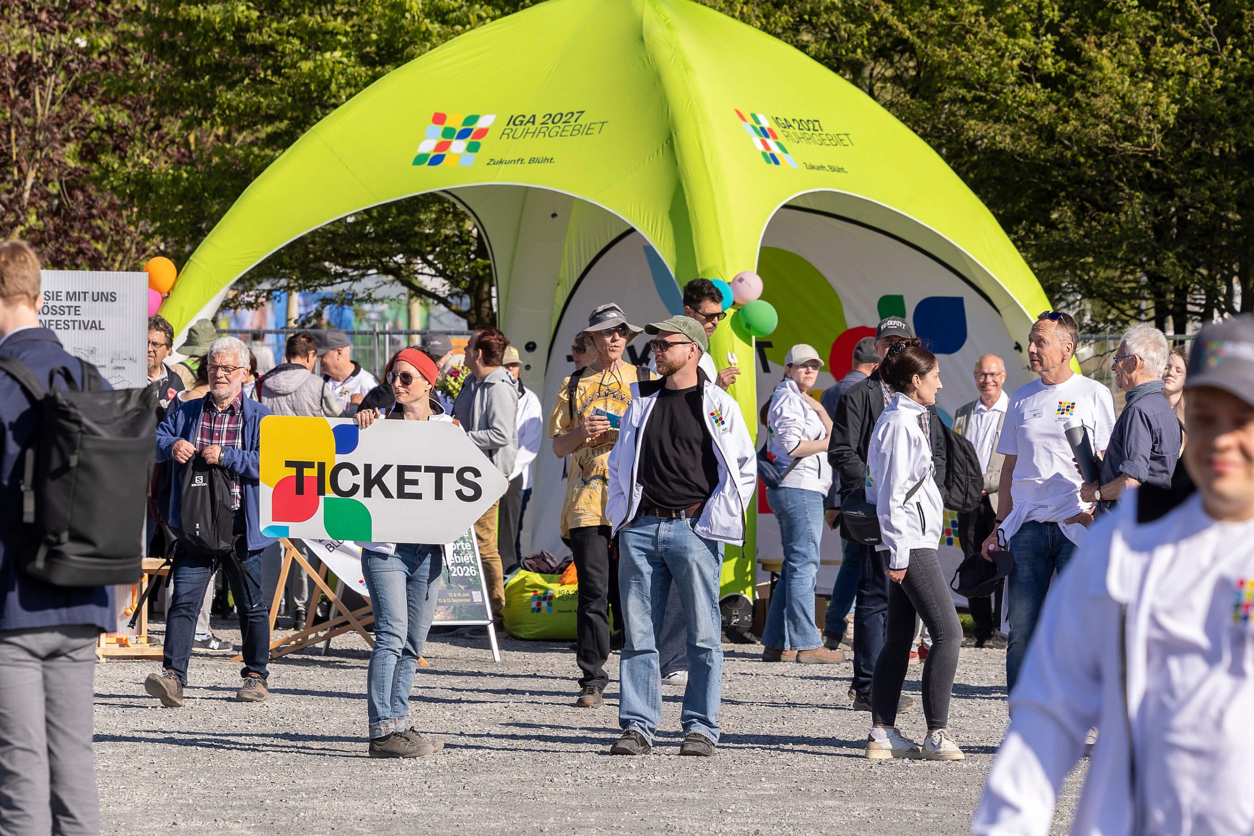 Hellgrünes Eventzelt mit dem Logo der IGA 2027 Ruhrgebiet, davor Besucherinnen und Besucher sowie eine Frau mit einem Wegweiser-Schild mit der Aufschrift „Tickets".