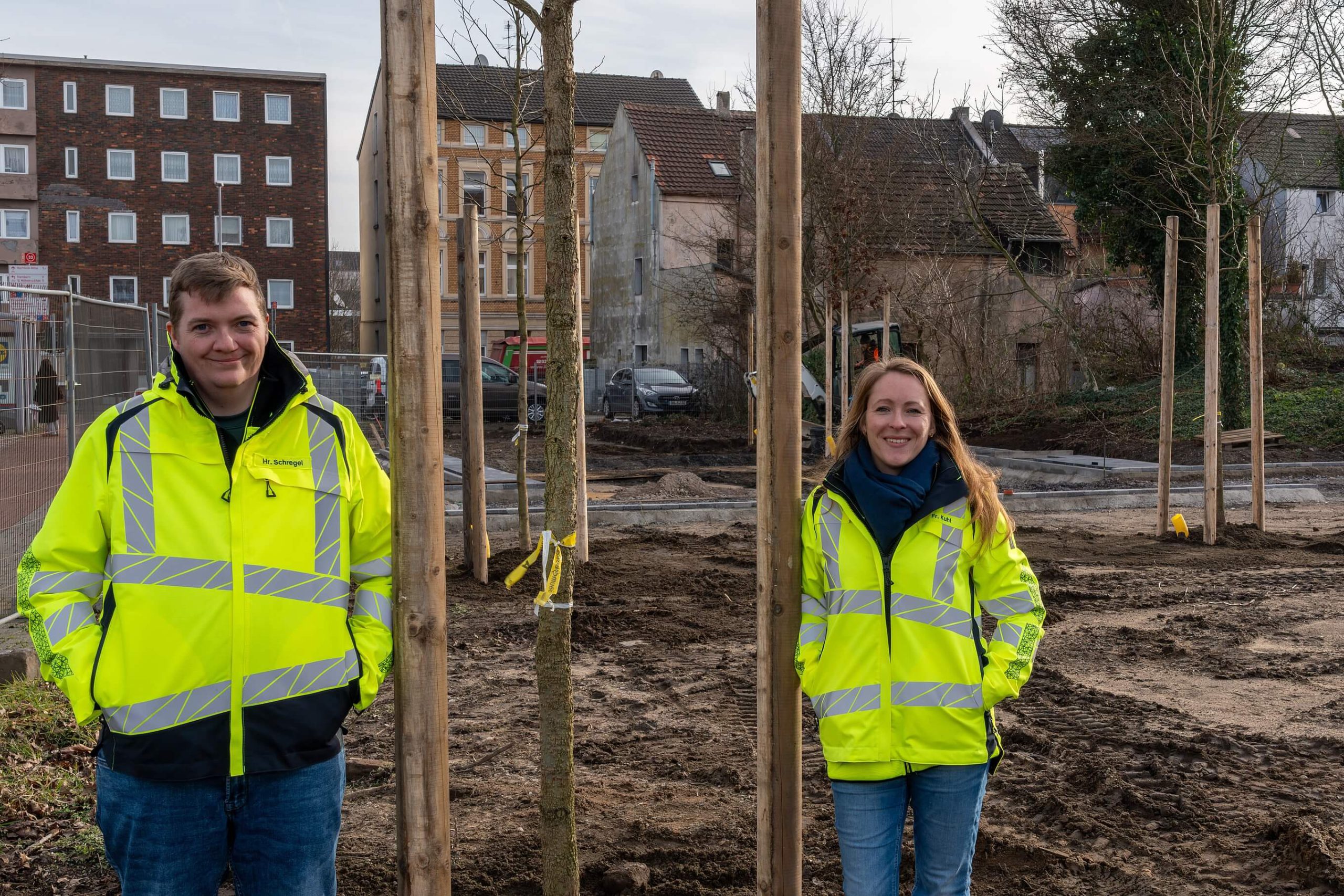 Zwei Beschäftigte in Warnjacken stehen zwischen frisch gepflanzten Bäumen auf der Baustelle am Grünen Ring Süd.