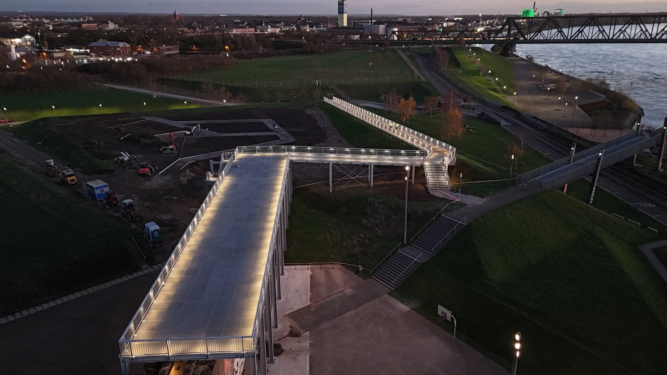 Luftaufnahme des beleuchteten Skywalks auf der Erzbunkerwand im RheinPark in der Abenddämmerung, im Hintergrund der Rhein und die Eisenbahnbrücke.