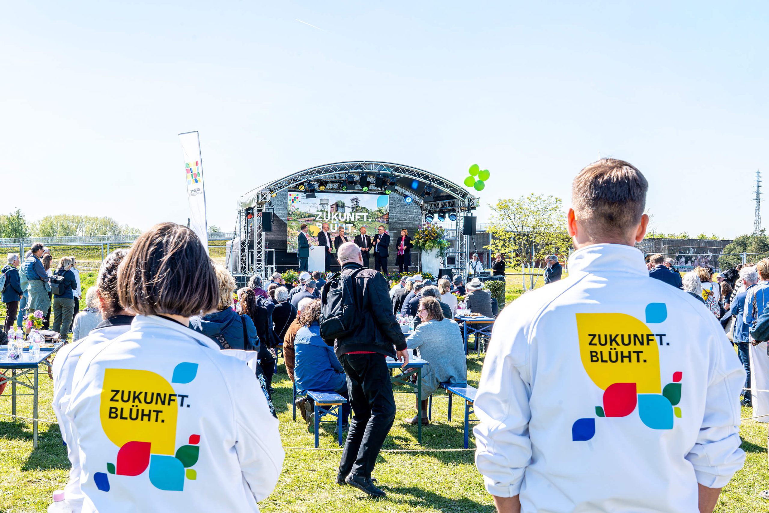 IGA-Auftaktveranstaltung im RheinPark: Publikum an Biertischen vor einer Bühne mit Podiumsgästen, im Vordergrund zwei Personen in Jacken mit der Aufschrift Zukunft. Blüht.