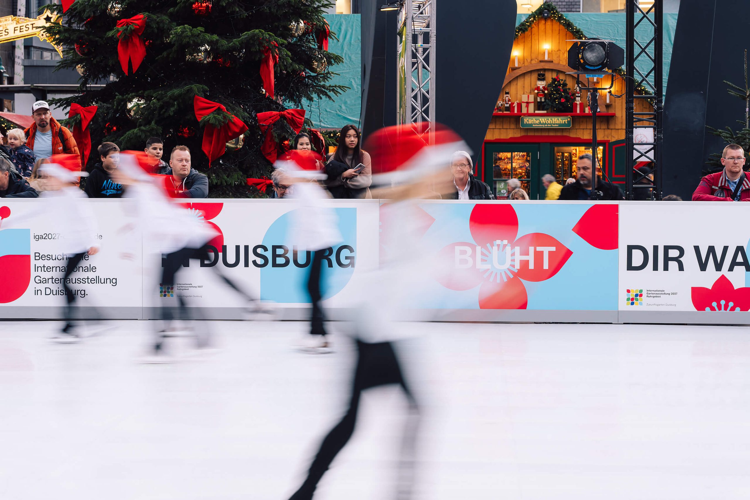 Eislaufende vor einer Bande mit IGA-Werbung und dem Schriftzug In Duisburg blüht dir was auf einer Eisbahn am Weihnachtsmarkt.