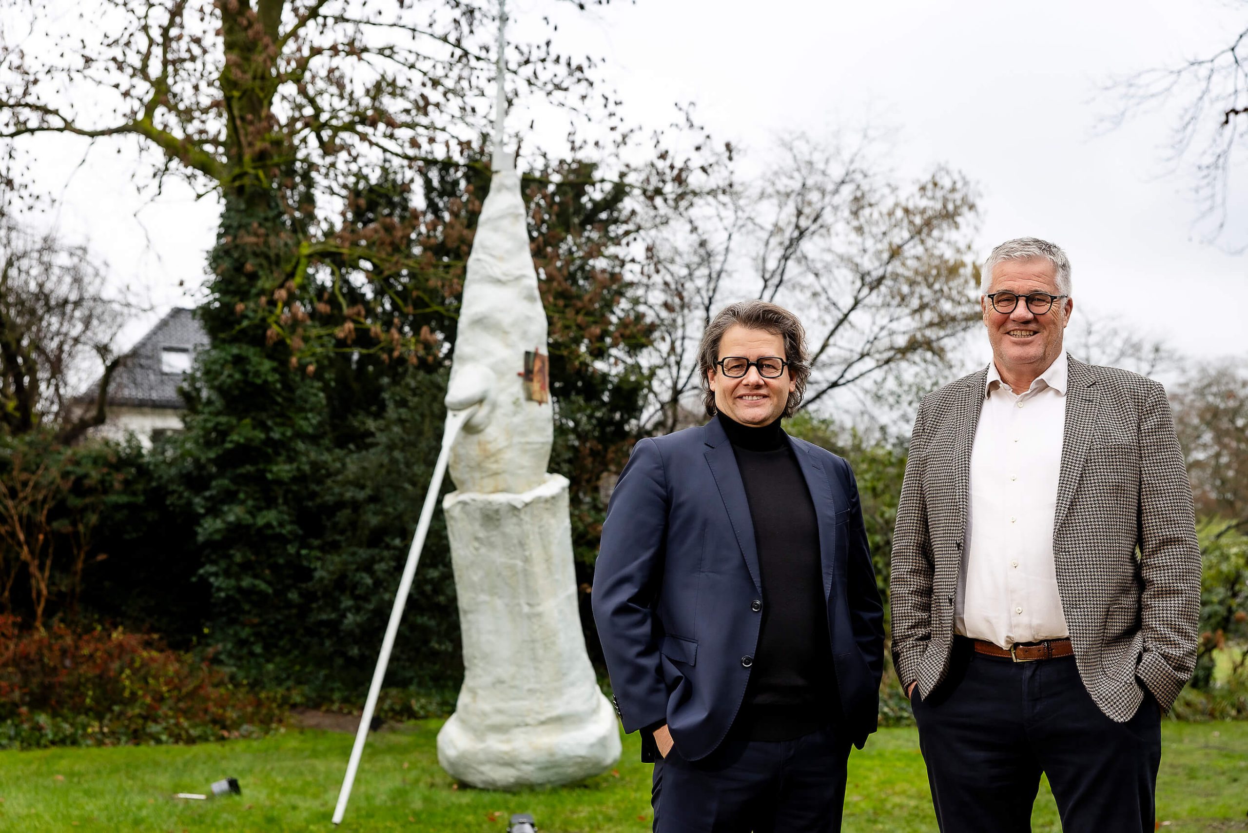 Zwei Männer stehen lächelnd in einem Garten neben einer weißen, turmartigen Skulptur mit Lanze. Foto: Anne Orthen.