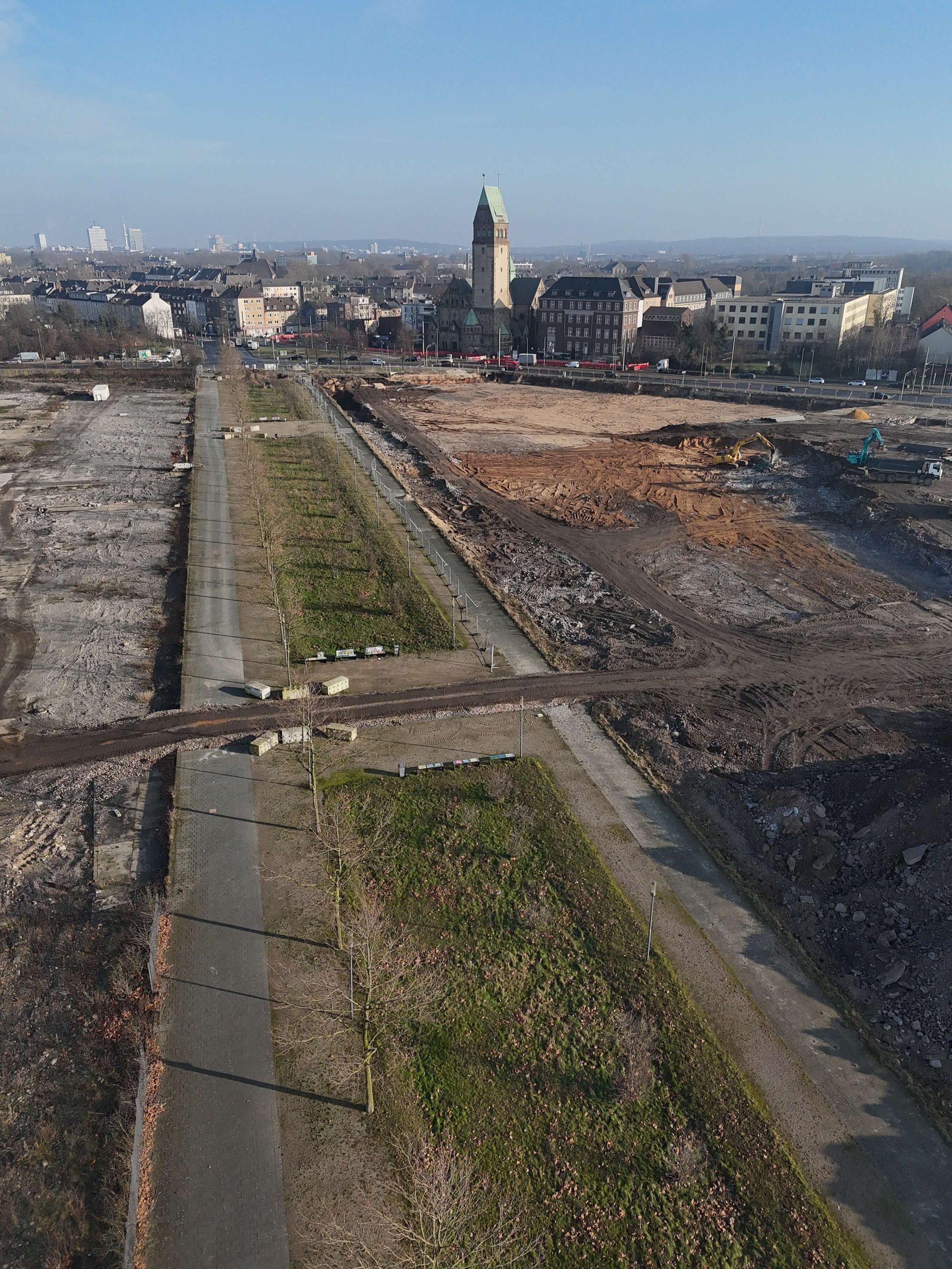 Luftaufnahme der Hochfeldallee im Bau: Baumreihe entlang eines bestehenden Wegs, rechts Erdarbeiten mit Baggern, hinten die Stadtkulisse mit Kirchturm.