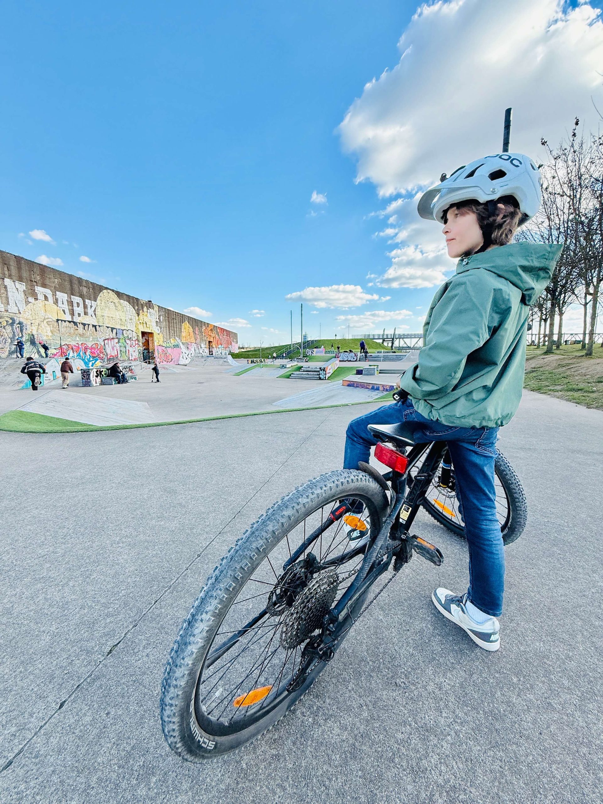 Kinder fahren mit Rollern über die Skateanlage im RheinPark, im Vordergrund ein Kind mit Helm, dahinter eine bunt bemalte Betonwand.