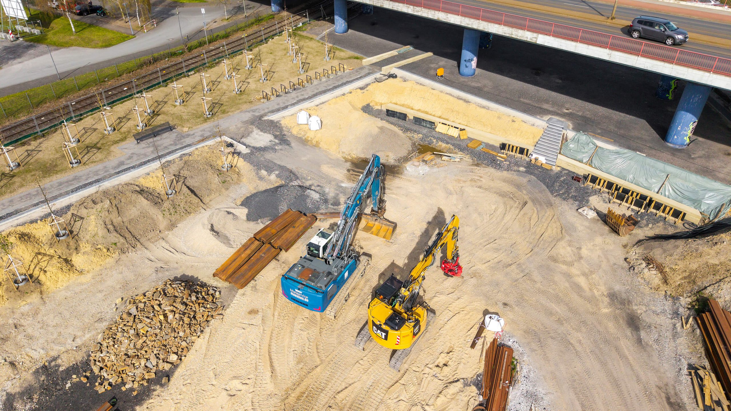 Luftaufnahme der Bauarbeiten an der Rampe zum Kultushafen: zwei Bagger auf der Erdbaustelle unter der Hochstraße, daneben junge Baumpflanzungen