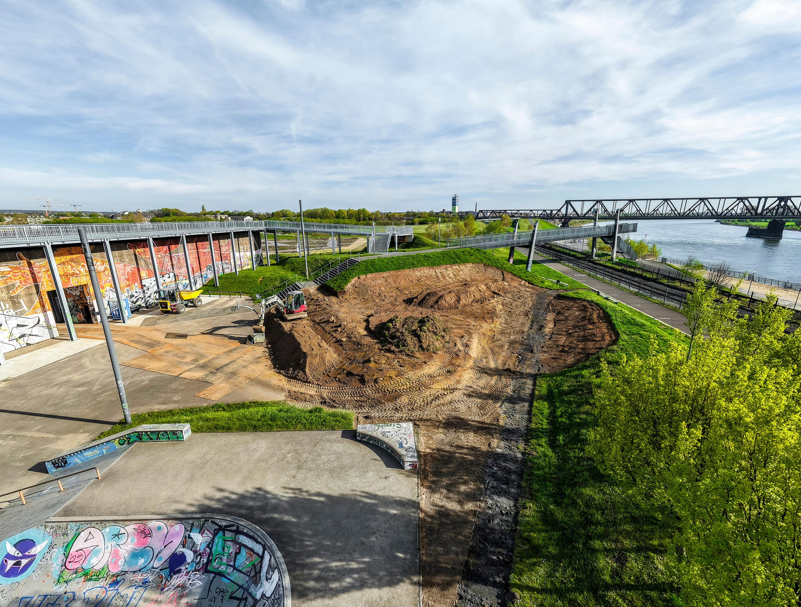Erweiterung der Skateanlage im RheinPark: Bagger modelliert eine Erdfläche neben der bestehenden Betonanlage, dahinter Rheinufer und Eisenbahnbrücke.
