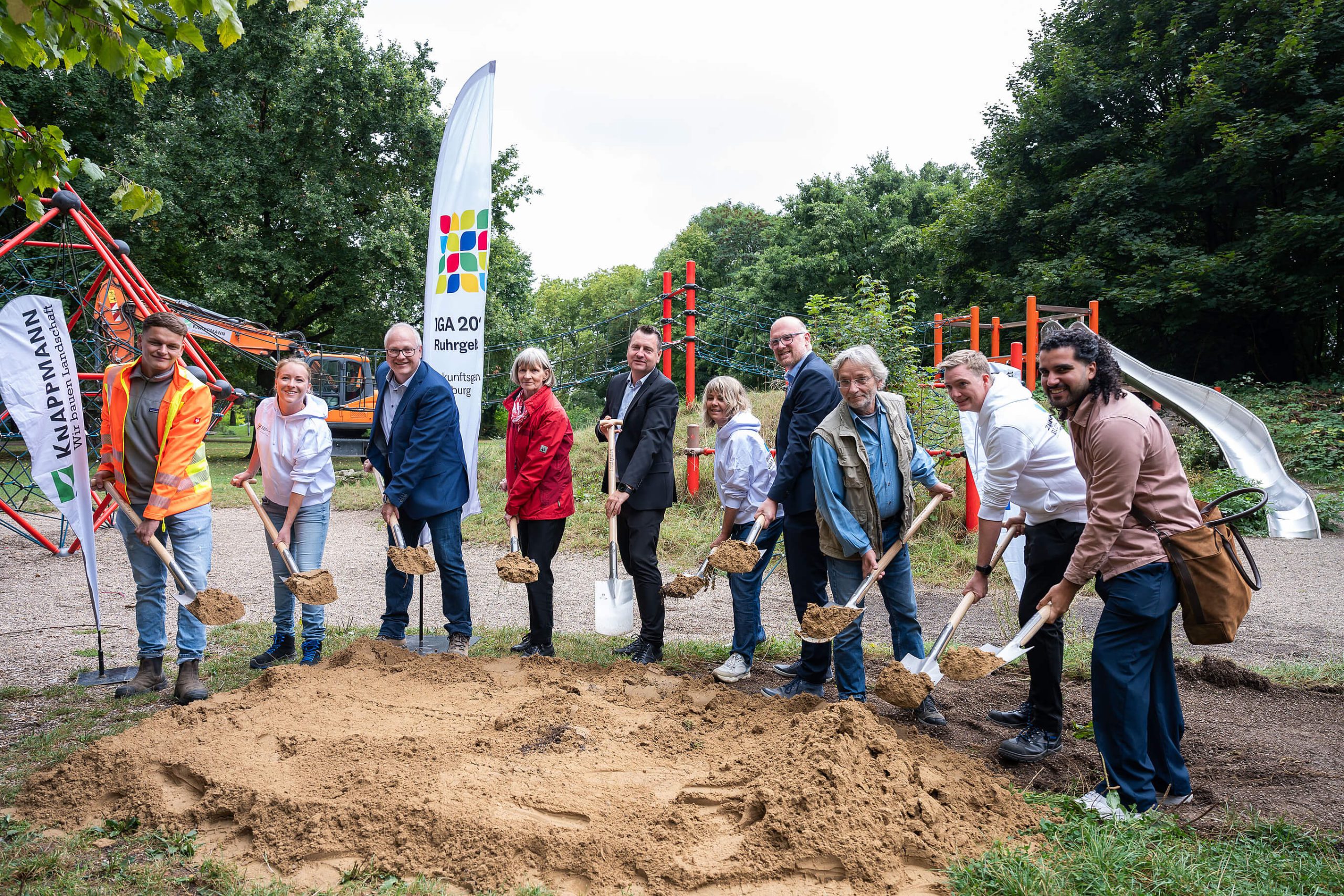 Spatenstich für den Grünen Ring: zehn Personen mit Spaten und Sandhaufen vor einem Spielplatz, daneben ein IGA-2027-Beachflag.