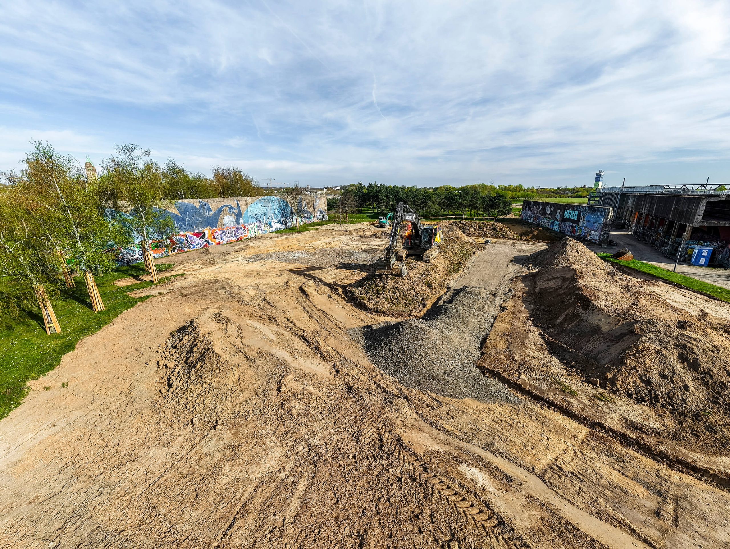 Erdarbeiten für den neuen Spielplatz im RheinPark: Bagger modelliert Hügel und Mulden vor der graffitibesprühten Bunkerwand.