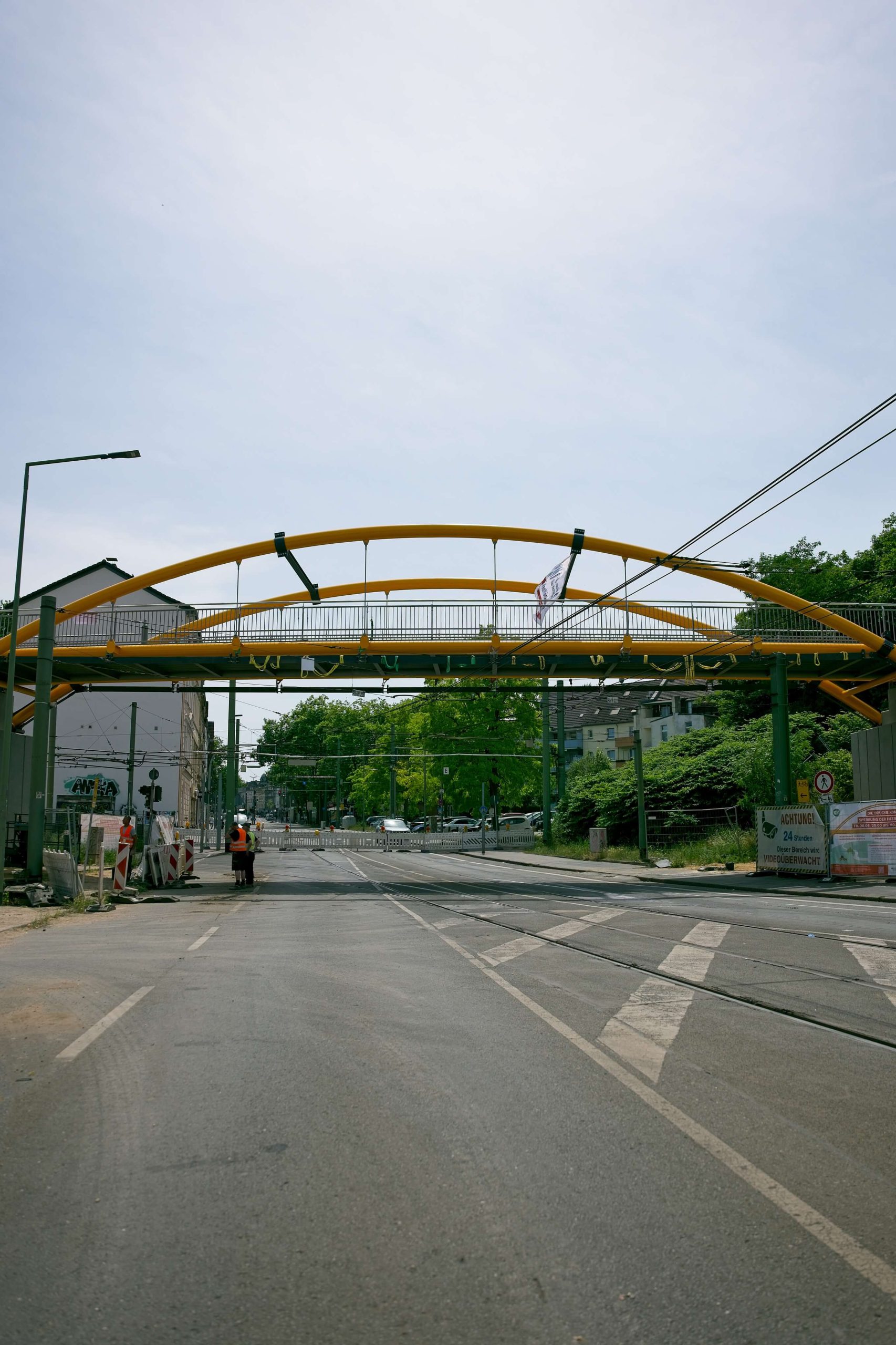 Neue gelbe Bogenbrücke über eine gesperrte Straße, davor Bauarbeiter und ein Hinweisschild zur Straßensperrung.