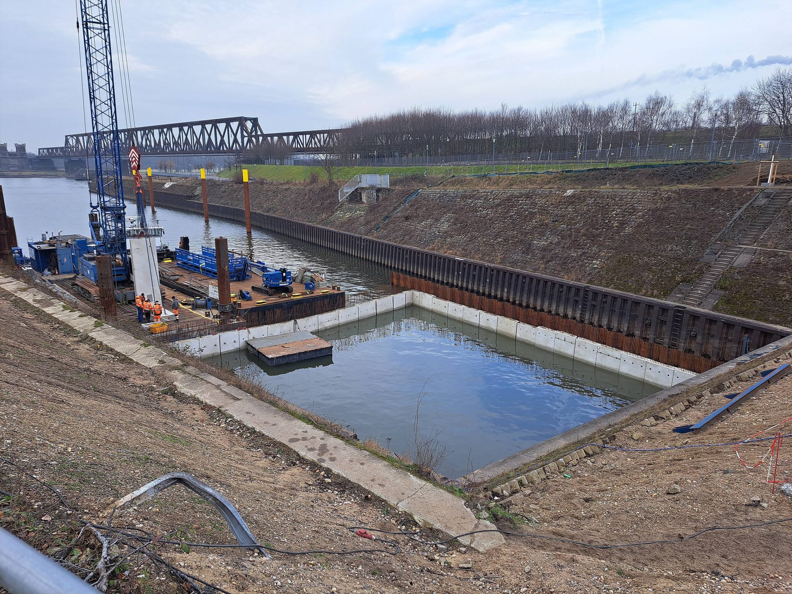 Verlängerung der Rampe ins Hafenbecken: Betonwände im eingespundeten Becken, daneben ein Ponton mit Kran und Bauarbeitern.