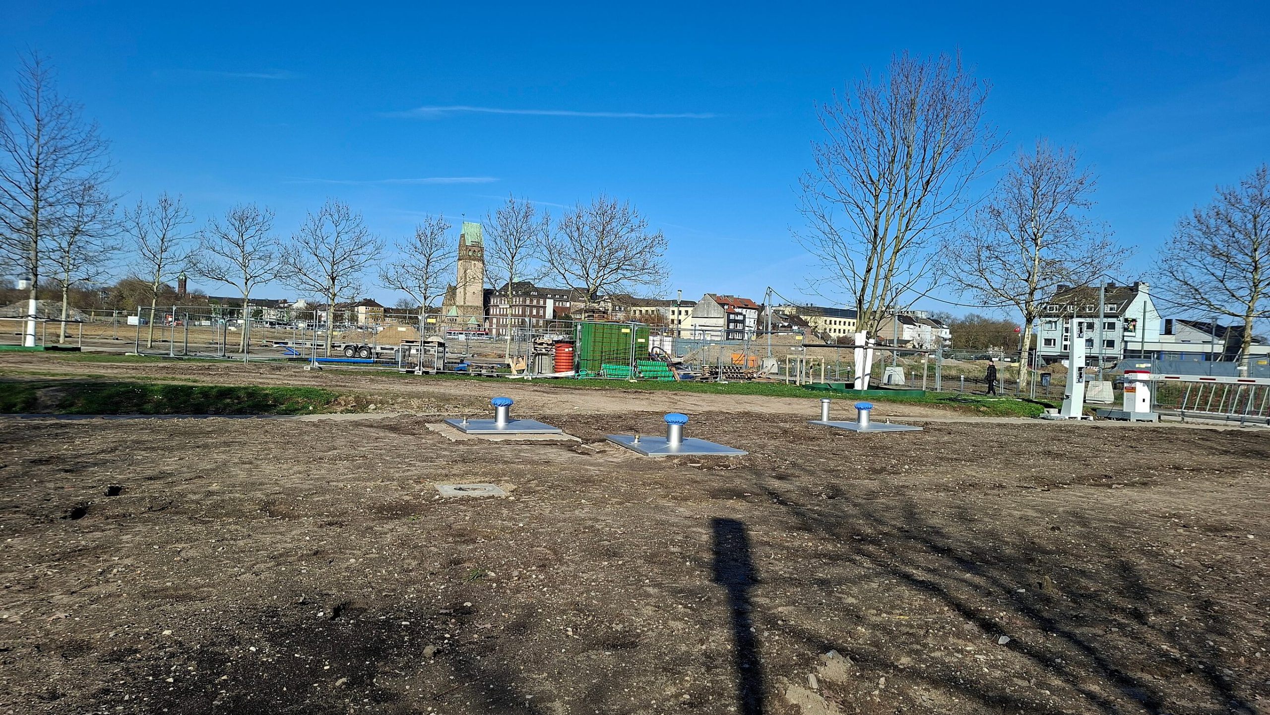 Baustelle der Zisterne im RheinPark: Schachtabdeckungen mit blauen Kappen auf einer Erdfläche, im Hintergrund die Stadtkulisse mit Kirchturm.