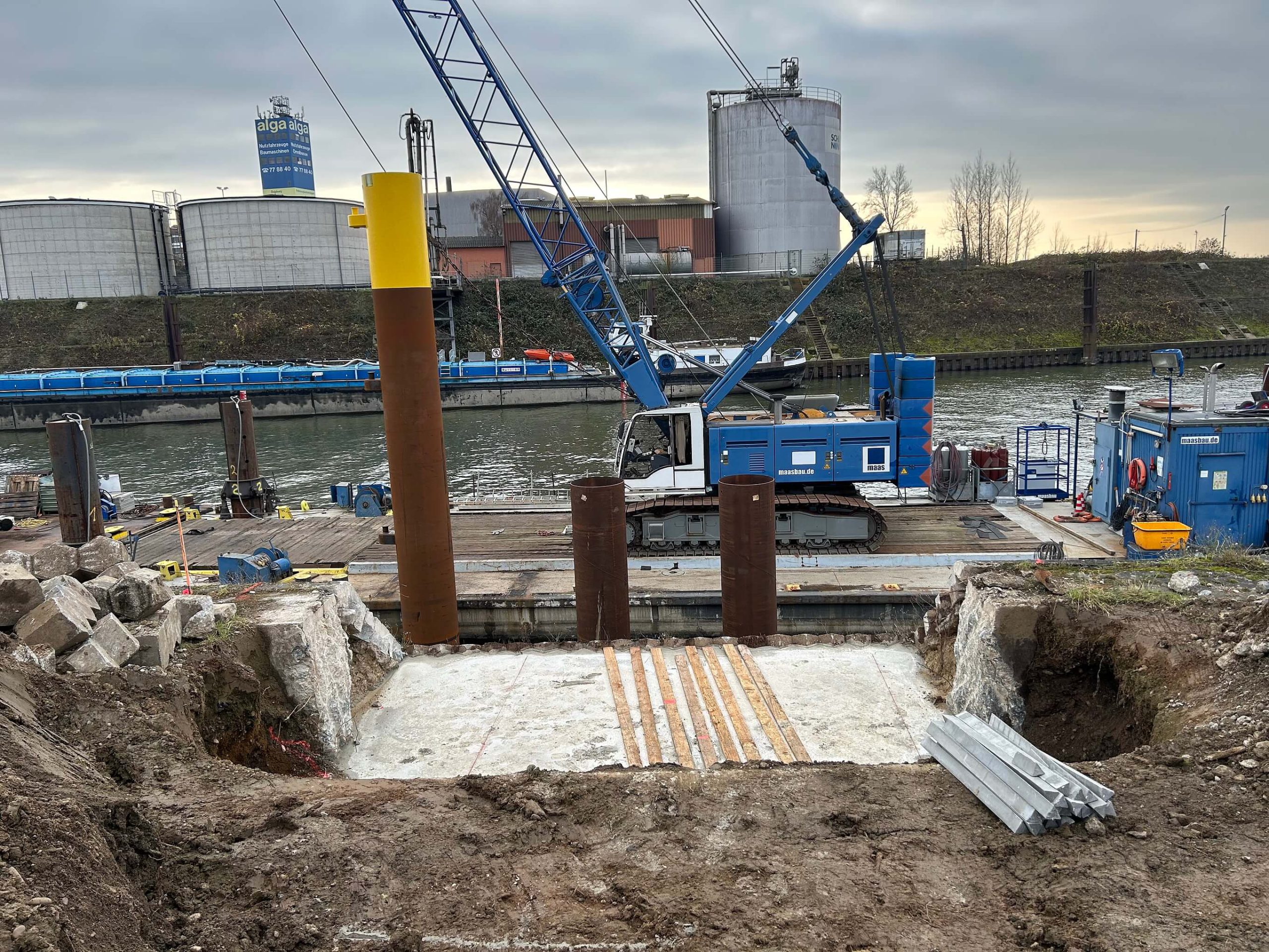 Gründungsarbeiten am Hafenbecken: Stahlrohre und Betonfundament in der Baugrube, dahinter ein Ponton mit blauem Kran.
