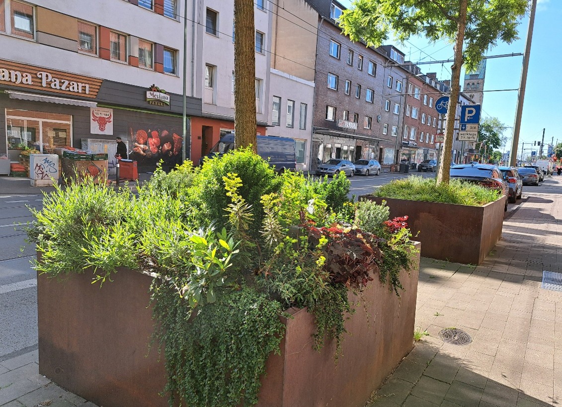 Bepflanzte Cortenstahl-Hochbeete auf dem Gehweg der Wanheimer Straße, dahinter Geschäfte und parkende Autos.