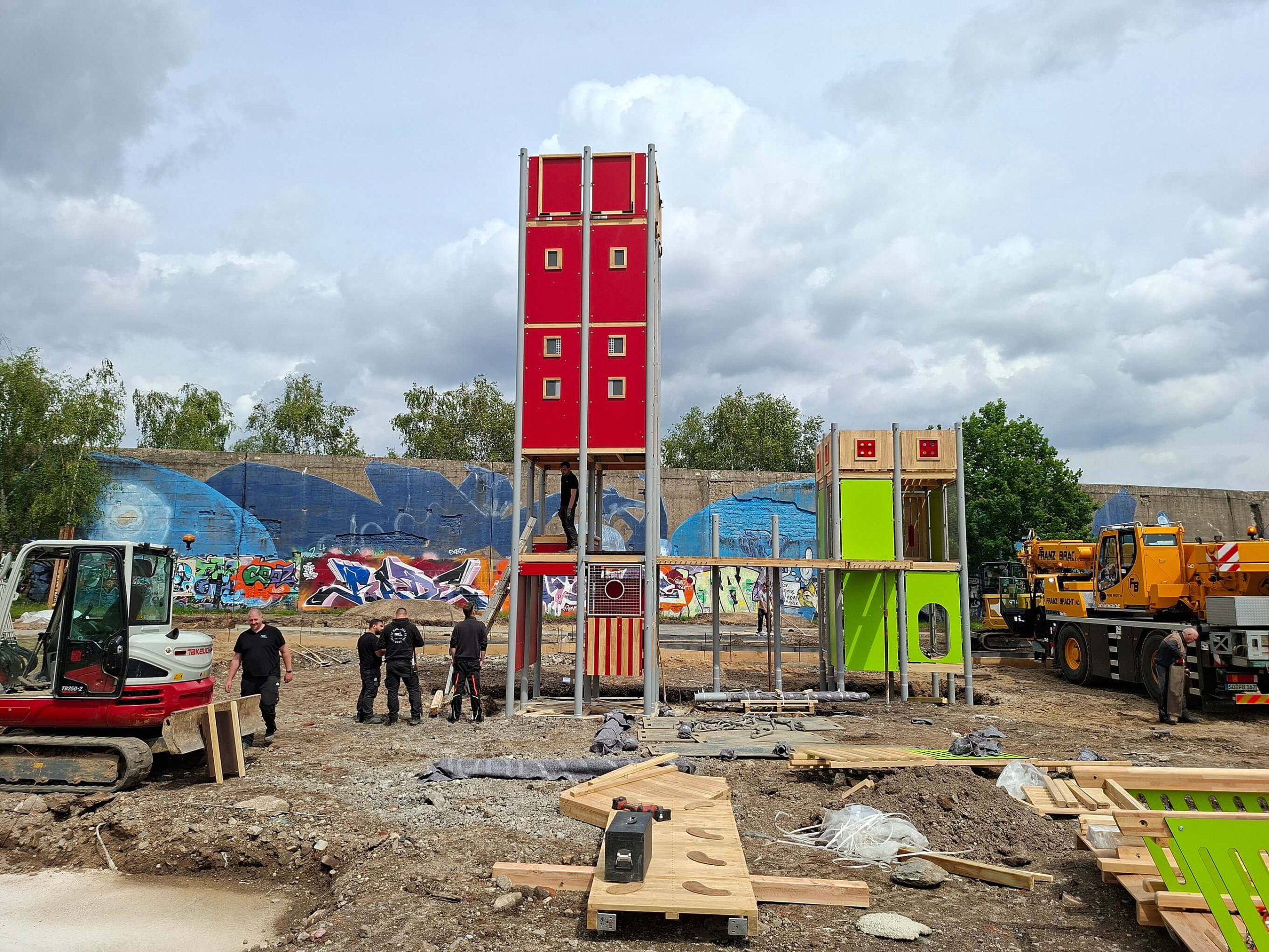 Aufbau des inklusiven Spielplatzes im RheinPark: Monteure errichten einen roten und einen grünen Spielturm vor einer graffitibesprühten Mauer