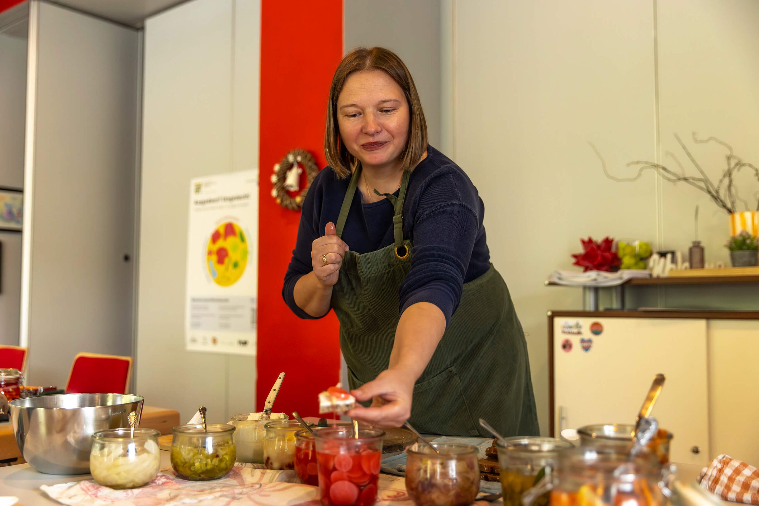 Eine Frau steht hinter einem Tisch mit verschiedenen Einmachgläsern und trägt eine Kochschürze. In der Hand hält sie ein Stück zubereitetes Essen.