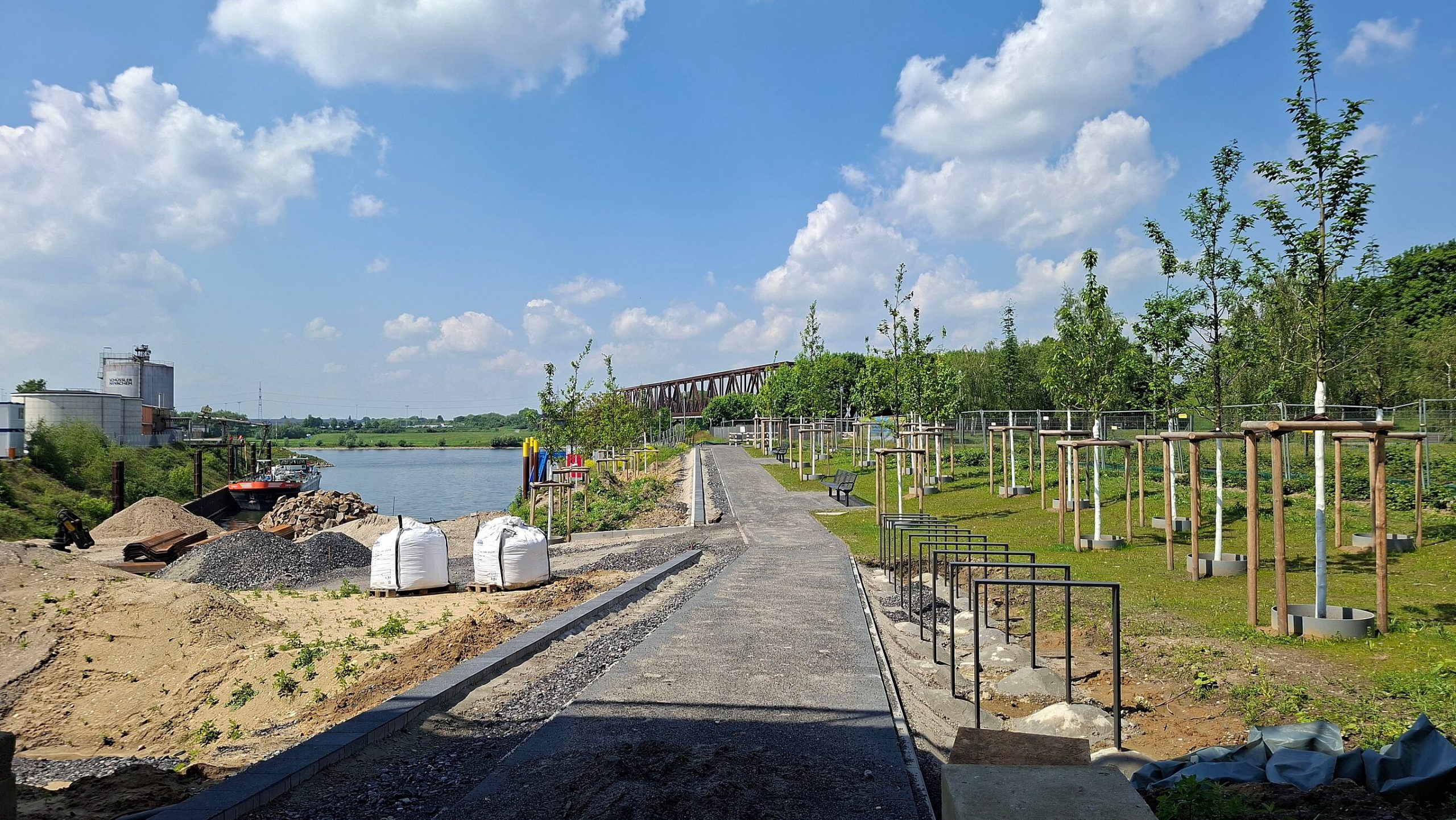 Neuer Uferweg am Kultushafen mit frisch gepflanzten Bäumen und Sitzbänken, links der Rhein, im Hintergrund die alte Eisenbahnbrücke