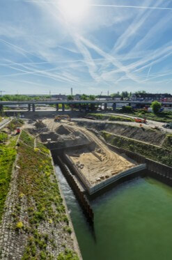 Blick auf die Bauarbeiten am Kultushafen. Im Hafenbecken ist der künftige Hafenbalkon mit Spundwänden eingefasst und teilweise verfüllt. Auf der Baustelle sind Bewehrungselemente und Baumaschinen zu sehen. Das angrenzende Gelände wird für die geplante Rampe modelliert.