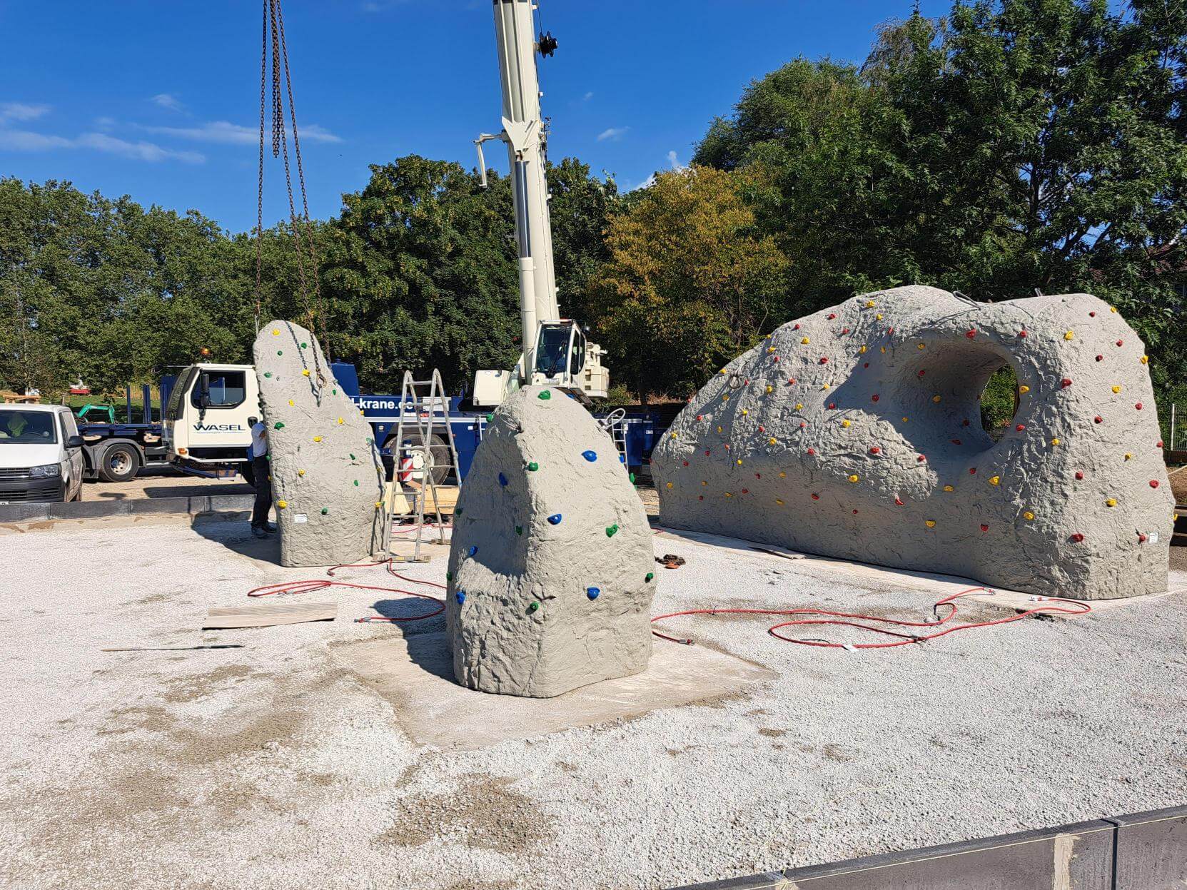 Drei große Kletterfelsen mit verschiedenen Elementen zum Bouldern stehen auf dem Gelände des Grünen Rings. Im Hintergrund ist ein Kran zu sehen.