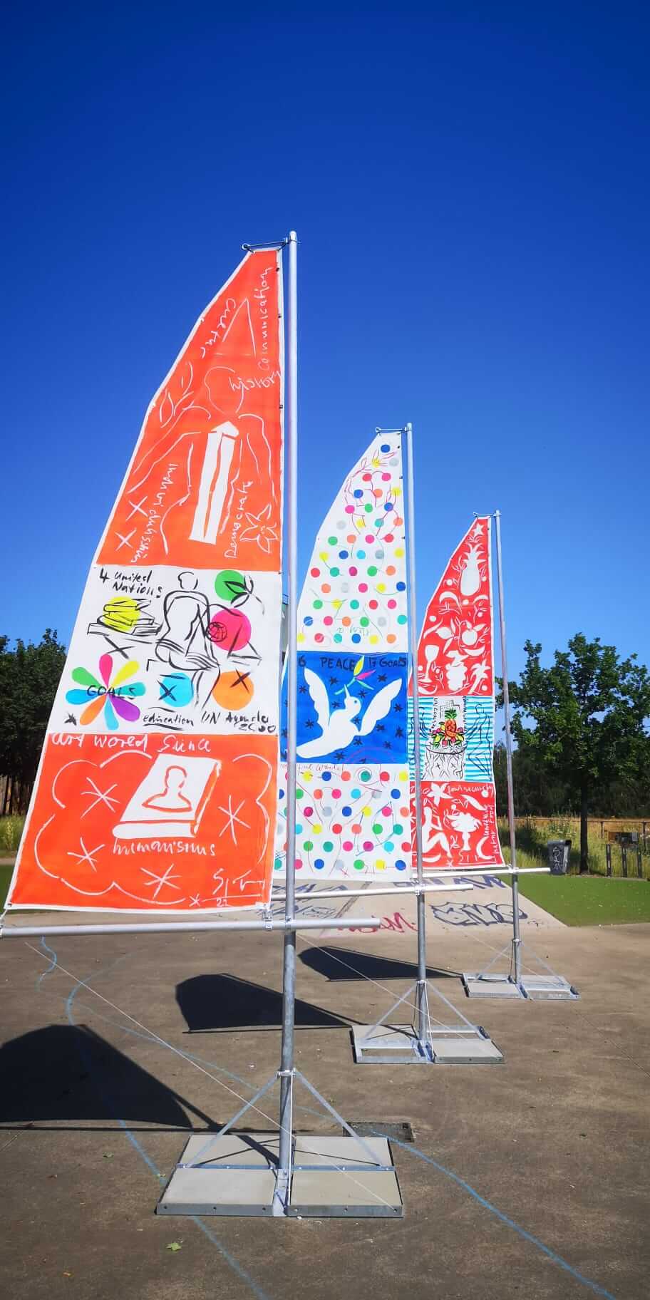 Drei gestaltete Beachflags stehen im Zukunftsgarten Duisburg. Die Fahnen sind mit Motiven und Gestaltungselementen des Internationalen Skulpturengartens der IGA 2027 versehen.