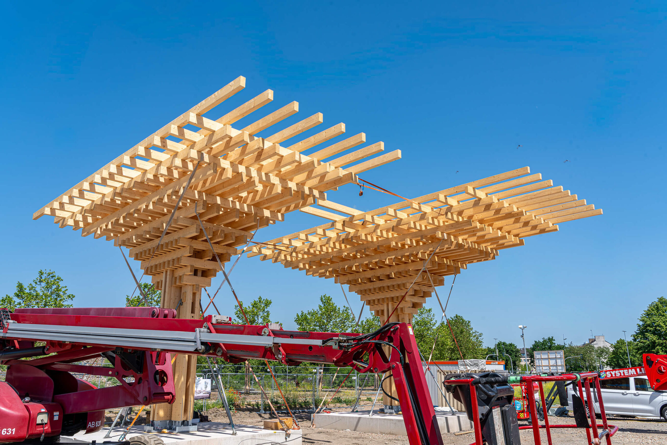 Zwei Elemente des Holzpavillons sind zu sehen. Daneben befindet sich ein Kran beziehungsweise Arbeitsgerät auf der Baustelle.
