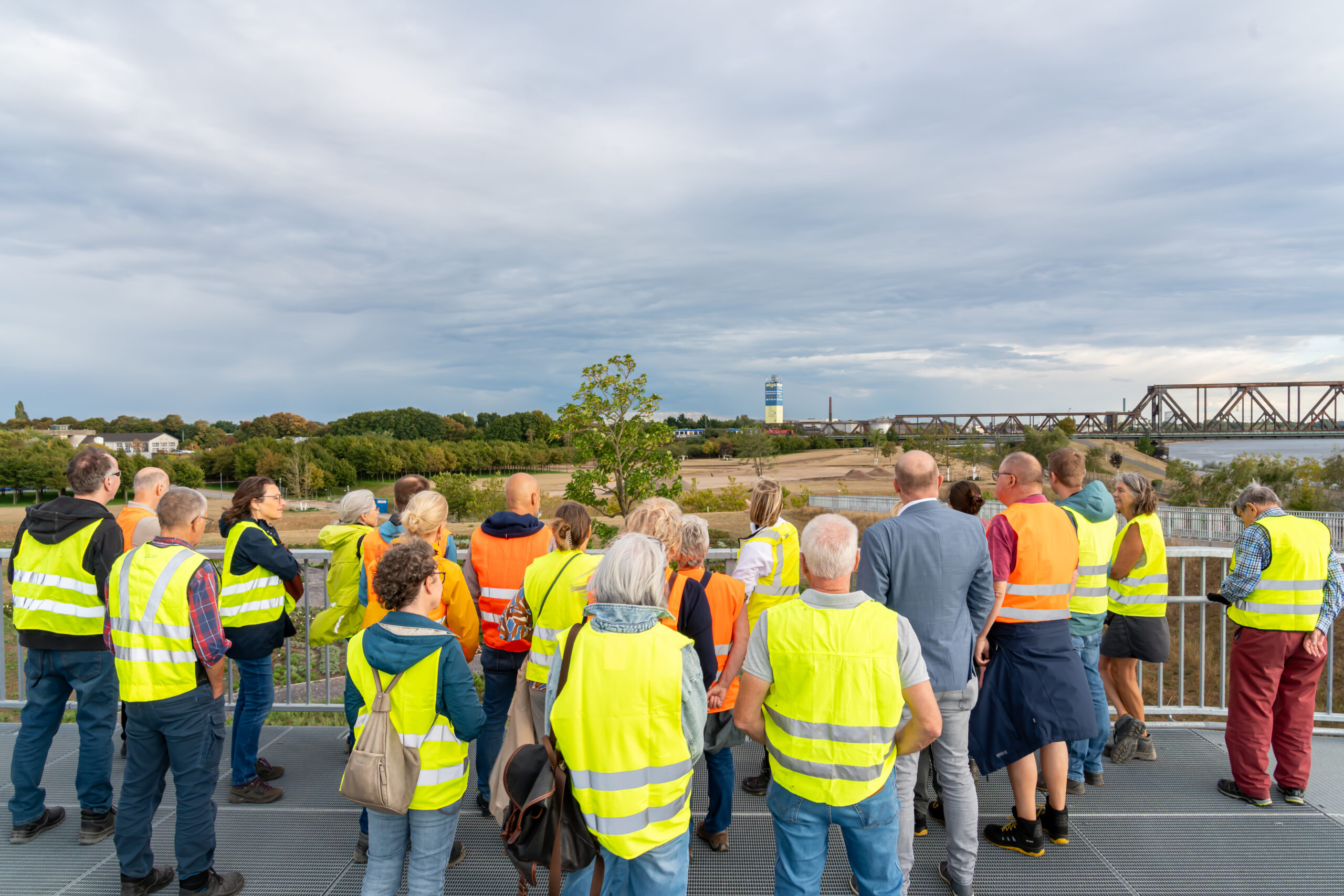 Menschen werden über das Gelände geführt. Alle tragen gelbe oder orangefarbene Warnwesten. Im Hintergrund ist eine Eisenbahnbrücke zu sehen.