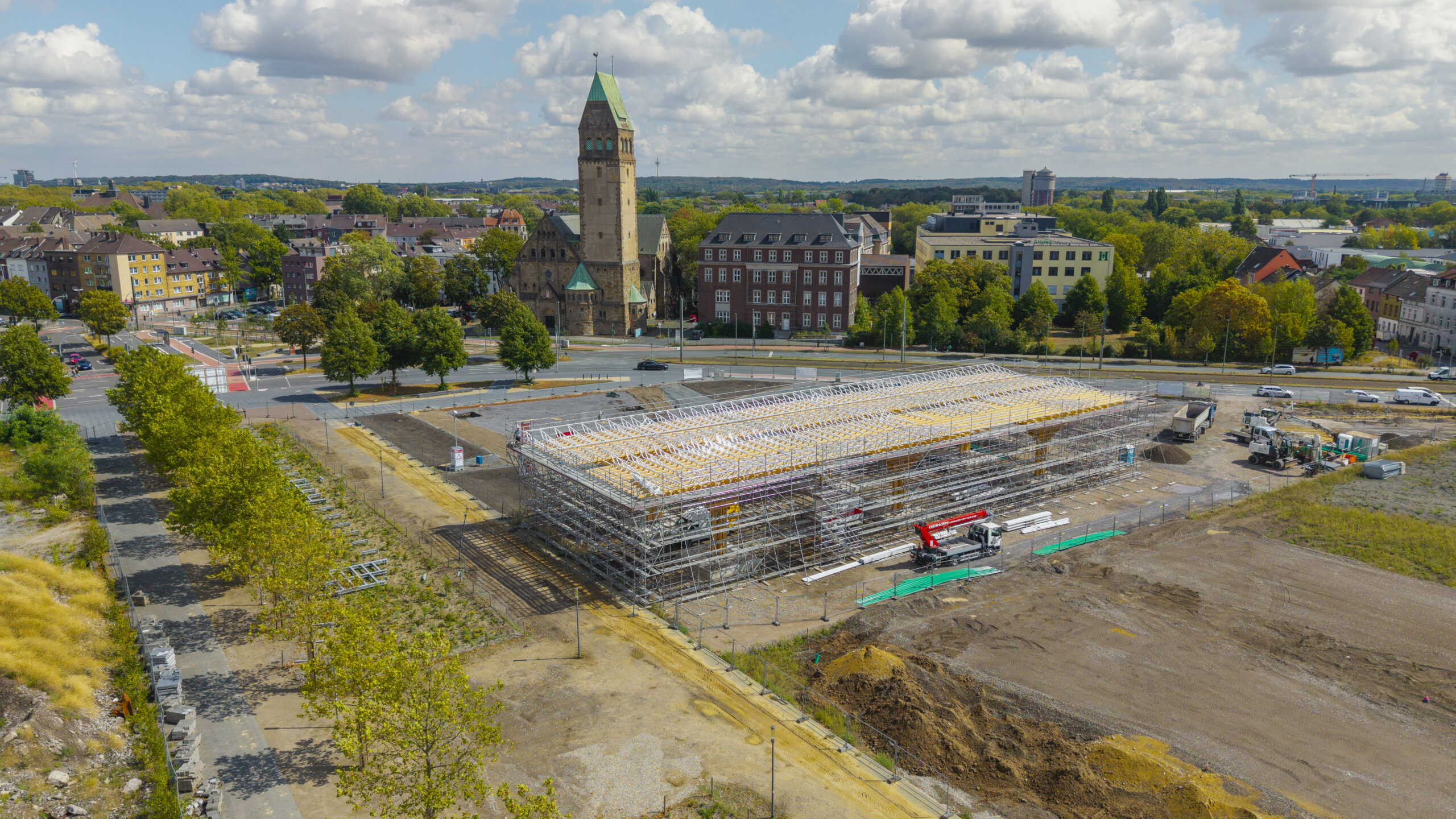 Der Pavillon ist aus der Luft zu sehen. Der Holzbau ist bereits fertiggestellt und wird derzeit von einem metallischen Gerüst gestützt. Rund um das Gebäude werden die angrenzenden Freiflächen vorbereitet.