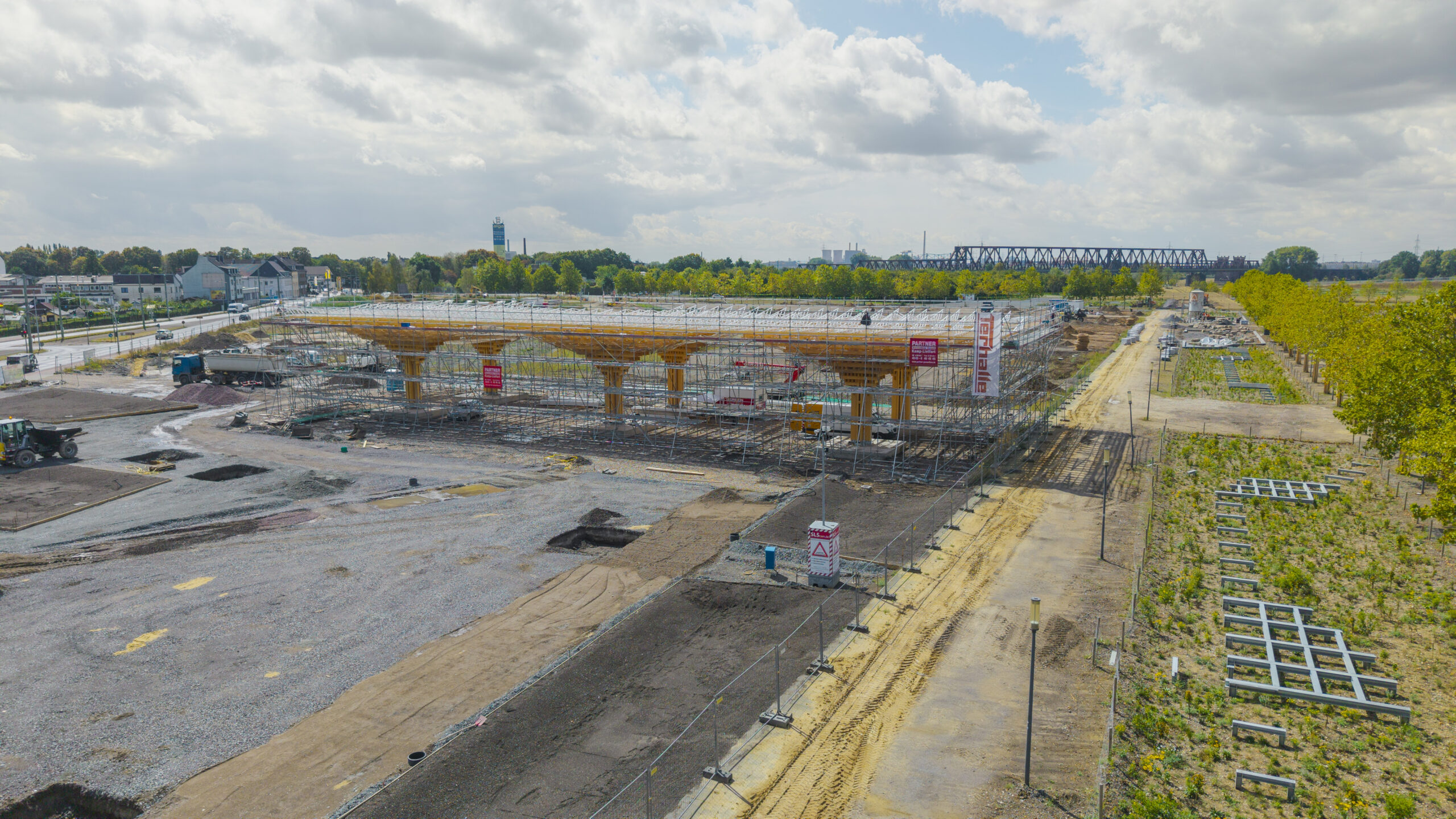 Der Pavillon ist aus der Luft zu sehen. Der Holzbau ist bereits fertiggestellt und wird derzeit von einem metallischen Gerüst gestützt. Rund um das Gebäude werden die angrenzenden Freiflächen vorbereitet.