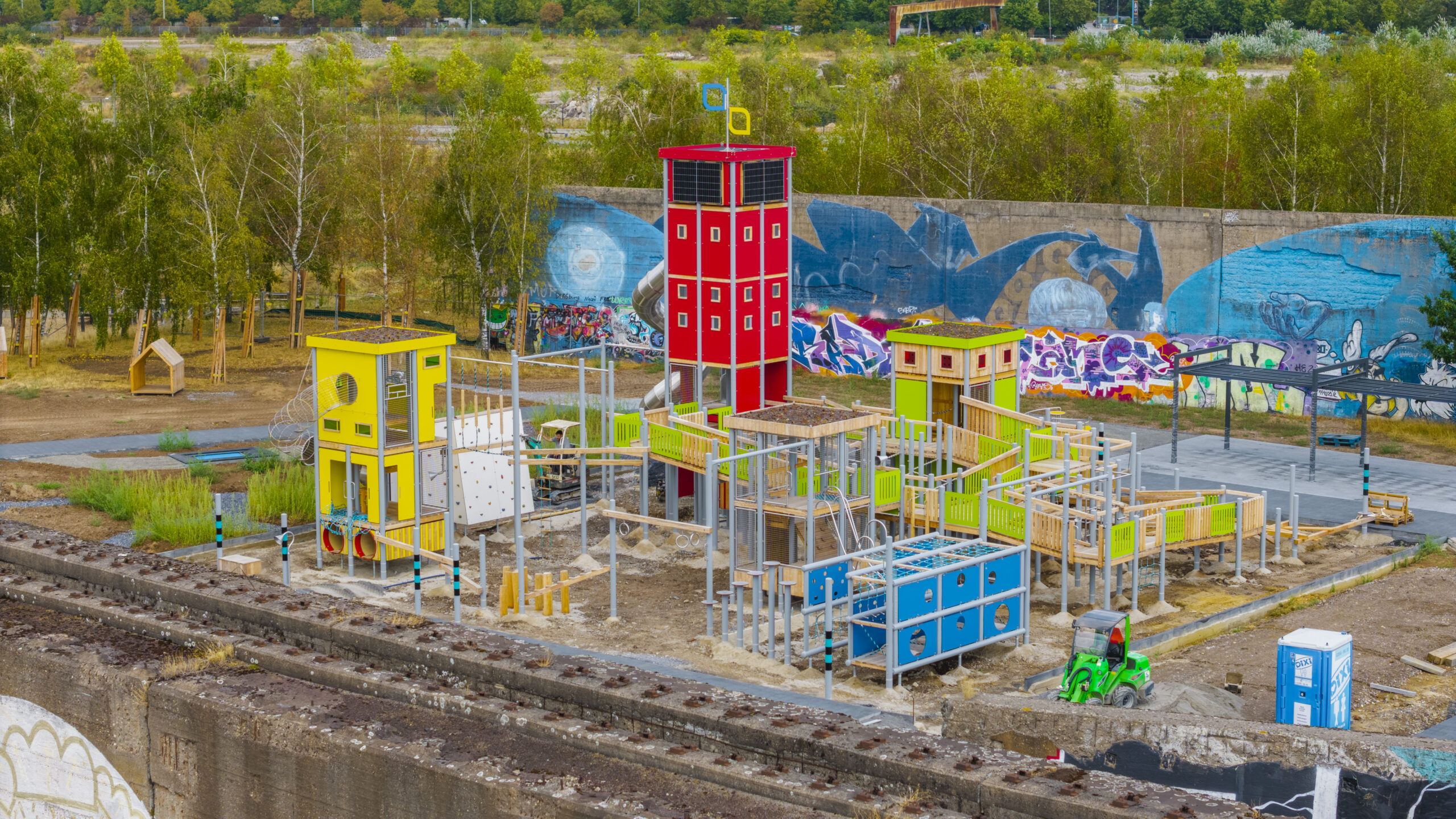 Der Aufbau des inklusiven Spielplatzes ist zu sehen. Die Anlage besteht aus Spiel- und Kletterelementen in Gelb, Rot, Grün und Blau. Im Mittelpunkt entsteht ein großes inklusives Spielgerät mit Möglichkeiten zum Klettern, Rutschen und Balancieren.